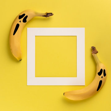 Halloween concept with two bananas with painted on them scary mask of fear and white frame with copy space for text on yellow background