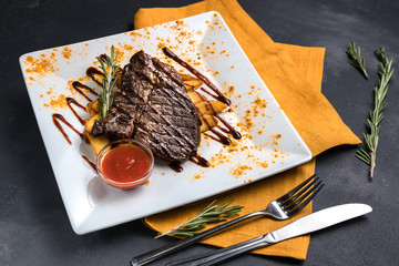 Grilled beef steak with potatoes, sauce and spices on white plate. Spoon, fork and orange tablecloth on dark gray texture background