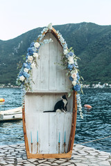 A wooden boat decorated with flowers as a wedding arch on the background of the sea. Preparing for the wedding. Inside is a curious cat.