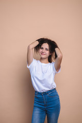 A girl looks at the camera and smiles, on a beige background, space for text. Portrait of a cute girl with clean skin who straightens her hair with her hands. Stylish girl in jeans and a white jacket