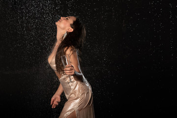 a brunette girl in a gold dress with a glass of champagne in her hand stands in the rain on a dark background