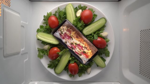Cell phone on plate with baked italian pizza on screen in microwave. Food blog concept. Top view of mobile phone with food footage on screen rotating in microwave oven.