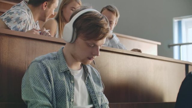 A male student in a University auditorium listens to music in white headphones during a break without paying attention to others. A lot of people in the audience are talking