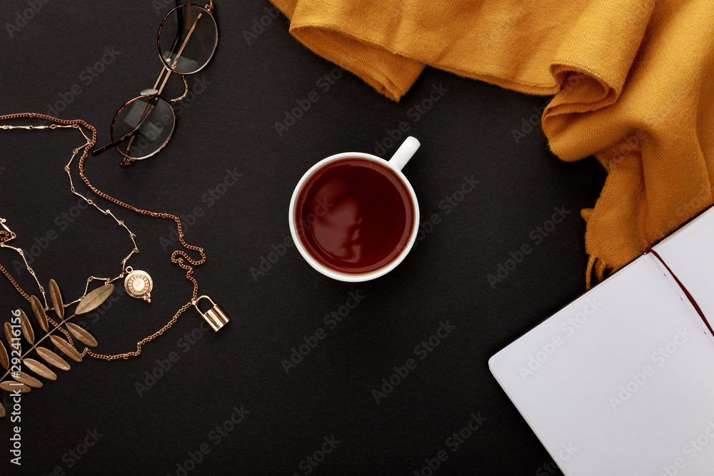 Wall mural top view of tea in mug near autumnal accessories and blank notebook on black background