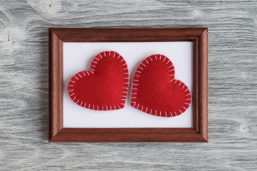 Two cute red felt handmade hearts lying on a brown frame on a gray wooden background. Top view. Flat lay
