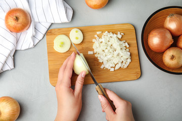 Woman cutting ripe onion on wooden board at grey table, top view
