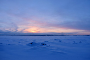 Obraz premium View on winter snowy river with snowbanks and blurred background with sunset, shore front and town chimney. Copy space