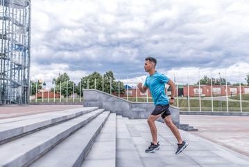 Athlete man runs in summer in city on morning run, background concrete steps, clouds, free space for text motivation. Active youth lifestyle, fitness workout man.