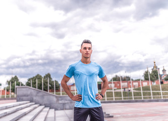 Confident and strong male athlete, summer city, trainer stares intently, background is blue clouds, free space for motivation text, fitness workout workout. Sportswear t-shirt, fitness bracelet.