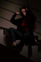 Young beautiful stylish woman wearing black jacket dress and hessian boots sitting on stairs in modern interior. Fashion style portrait with hard light