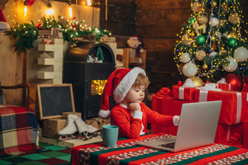 Christmas shopping concept. Gifts service. Little genius. Santa little helper. Little boy santa hat and costume having fun. Boy child with laptop near christmas tree. Buy christmas gifts online