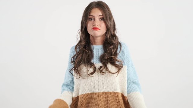Doubtful brunette girl knitted sweater throwing up hands guiltily looking in camera over white background. I don't know expression