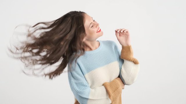 Attractive charming brunette girl in cozy sweater joyfully waving head and playing with hair on camera isolated