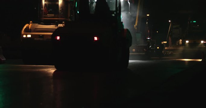 Road rollers silhouette working at night city on new road construction site. Truck laying fresh asphalt in town. Team of workers put hot asphalt on a street. Steam rising from hot asphalt surface.