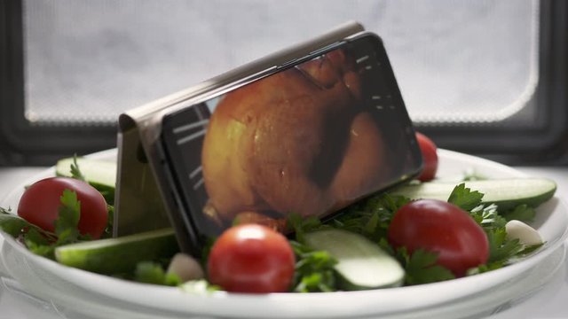 Cellphone with Thanksgiving or Christmas roasted turkey on screen in microwave. Plate with salad and mobile phone with cooking food footage on screen rotating in microwave.