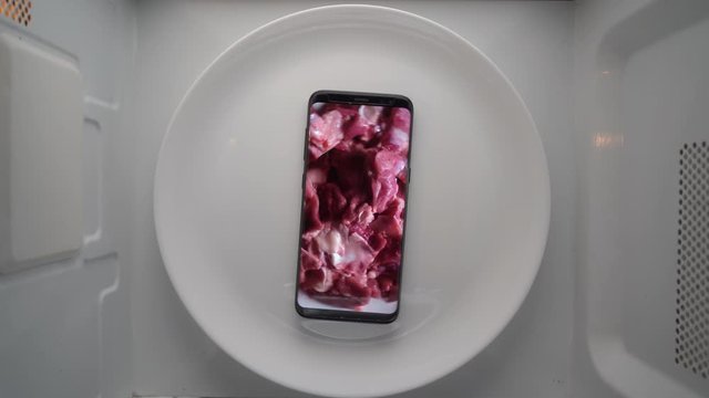 Cell phone on a plate with raw turkey meat pieces on screen in microwave. Food blog concept. Top view of mobile phone with food footage on screen rotating in microwave oven.