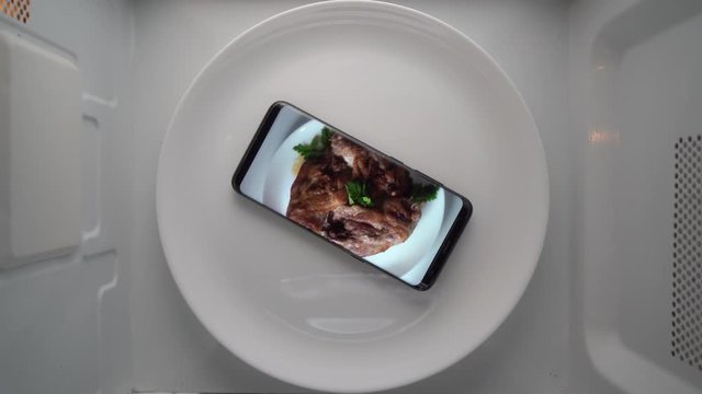 Cell phone on plate with fried chicken legs on screen in microwave. Food blog concept. Top view of mobile phone with food footage on screen rotating in microwave oven.
