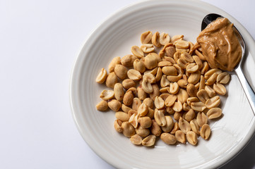 A close up top down view of a white plate on a white background holding peanuts and a spoonful of peanut butter.