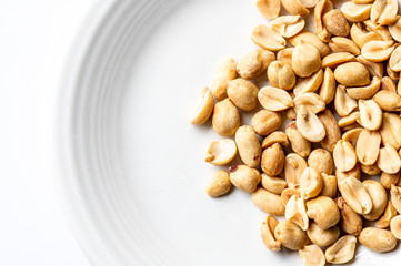 Close up of whole and half roasted peanuts on a white plate sitting on a white background.
