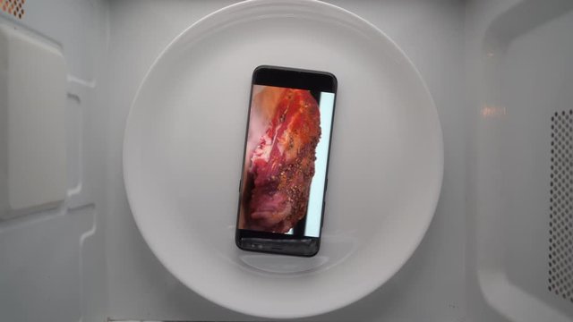 Cell phone on plate with fat pork meat roasting on screen in microwave. Food blog concept. Top view of mobile phone with food footage on screen rotating in microwave oven.