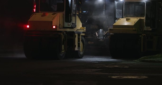 Road rollers silhouette working at night city on new road construction site. Truck laying fresh asphalt in town. Team of workers put hot asphalt on a street. Steam rising from hot asphalt surface.