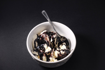 White bowl with serving of vanilla ice cream covered with melted chocolate topping along with a spoon on dark background