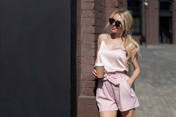 beautiful blonde girl holds glass coffee in her hand and stands near an office brick building during break. Copy space for coffee shop advertisement