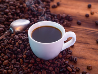 Coffee beans pilled on brown table with teaspoon and white coffee cup full of aromatic espresso. Texture of wooden desk. Selective soft focus. Blurred background