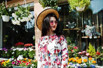 Fototapeta na wymiar Summer portrait of brunette girl in pink glasses and hat against flowers shop.