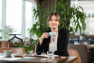 Young woman drinking fragrant coffee. Female holding cup. Girl Sitting in coffee shop at wooden table. large Windows, panorama of the city