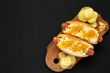 Obraz na płótnie Canvas Homemade colombian hot dogs with pineapple sauce, chips and mayo ketchup on a rustic wooden board on a black surface, overhead view. Top view, flat lay, from above. Copy space.
