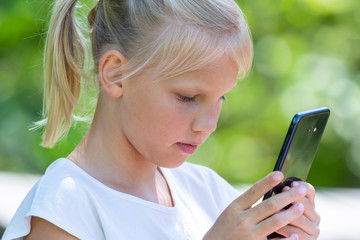 Young blond haired girl on black mobile phone in nature and park. Constant connection of the millennial generation. Screen time. Parenting concerns.