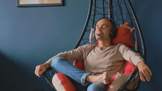 Young man in headphones sitting in hanging chair cocoon and relax. Blue wall with 4K slow motion video.