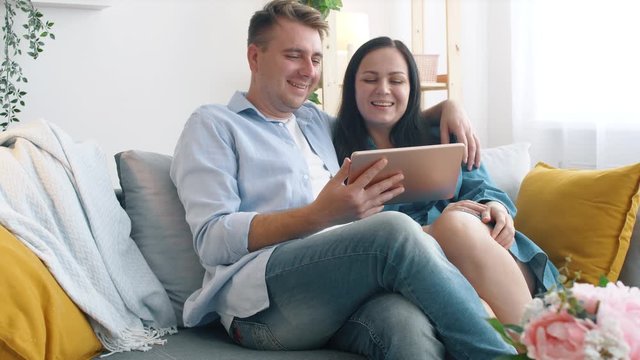 Young couple watching video on tablet while sitting on sofa in living room. Happy people relaxing at home using a tablet. Lifestyle concept.