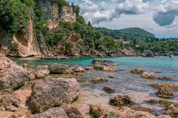 Seascape – lagoon with turquoise water, mountain with cliffs, green trees, bushes, rocks in a blue water and clouds on the sky. Corfu Island, Greece. 