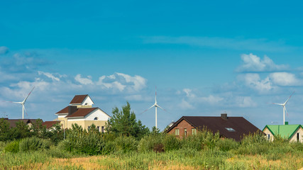 Fototapeta na wymiar Wind turbines behind houses in residential area on blue sky background. Clean and renewable energy concept