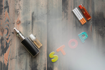 Flat lay of e-cigarette, e-liquid, tobacco cigarettes with lighter and letters stop on wooden background, covered smoke