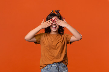 Smiling brunette woman in a t-shirt and beautiful headband hiding behind palms of hands standing isolated over orange background. Concept of positive surprise