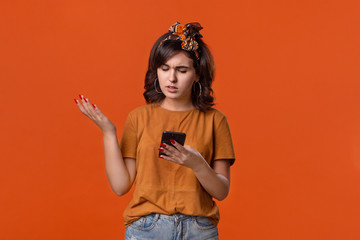 Pretty brunette woman in a t-shirt and beautiful headband is displeased checking smartphone for notifications isolated over orange background. Daily communication