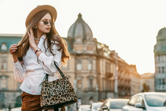 Outdoor fashion portrait of elegant, luxury woman wearing beige hat, sunglasses, trendy white shirt, brown wrist watch, holding animal, leopard print bag, posing in street. Copy, empty space for text