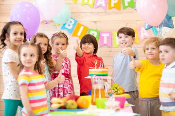 Fototapeta premium Children celebrating birthday party. Kids stand arround festive table and show thumbs up