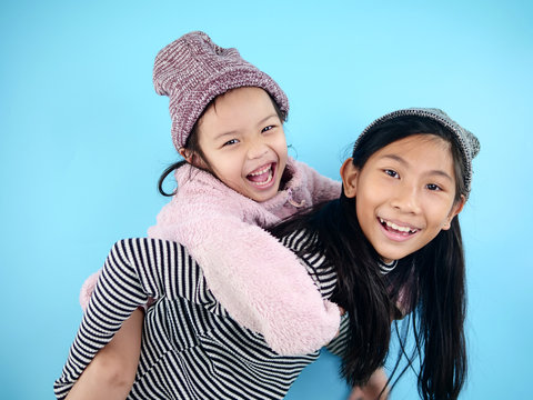 Laughing Asian Girl Enjoying Riding On Her Older Sister Piggyback Isolated On Blue Background.