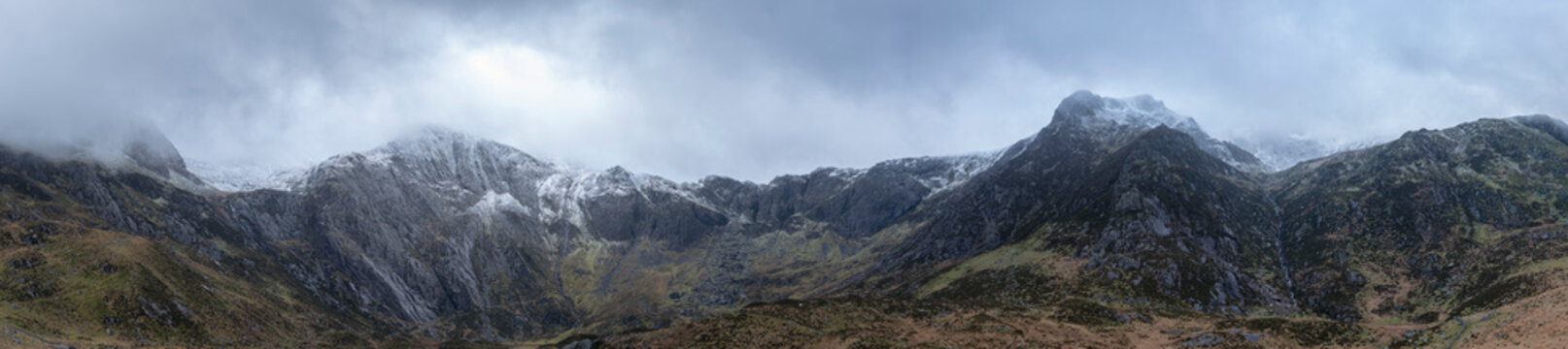 Stunning dramatic panoramic landscape image of snowcapped Glyders mountain range in Snowdonia during Winter with menacing low cloudshanging at the mountain peaks