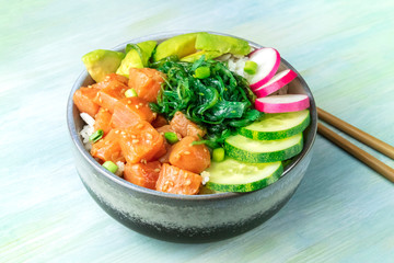 A bowl of salmon poke with avocado, wakame and cucumber on a teal blue background