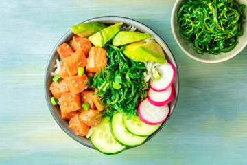 A bowl of salmon poke with avocado, wakame and cucumber, shot from the top on a teal blue background