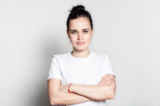 Pretty, Confident Young Woman Stands With Folded Arms, Stares Intently At The Camera And Smirks, On A White Background.