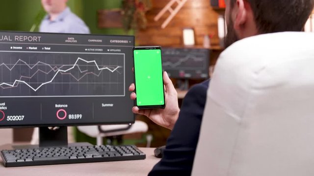 Businessman in modern workplace holding phone with green screen. Graph on PC monitor.