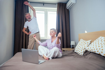 blonde short haircut woman meditating with laptop yoga video teacher in nightwear and elderly man doing stretching hands,spine in day light bedroom apartment