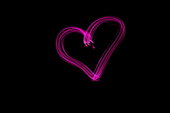Long exposure photograph of a heart shape outline in pink neon colour in an abstract swirl, parallel lines pattern against a black background. Light painting photography.
