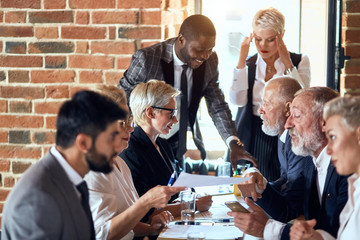 Group of businessmen wear suits sit at table in office and discuss new project, smile. caucasian blond woman and african man stand, behind open window. Everyone work